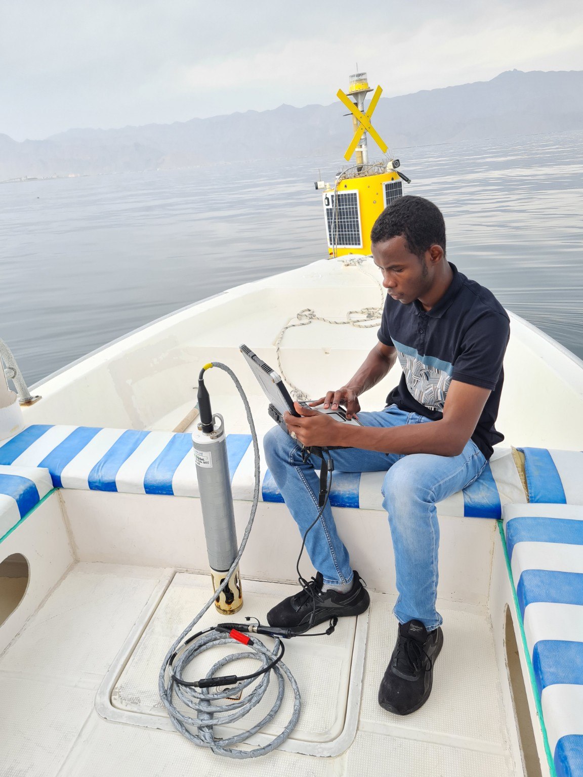 Husam working with a field computer and water-quality instrument on a boat, with a monitoring buoy behind him